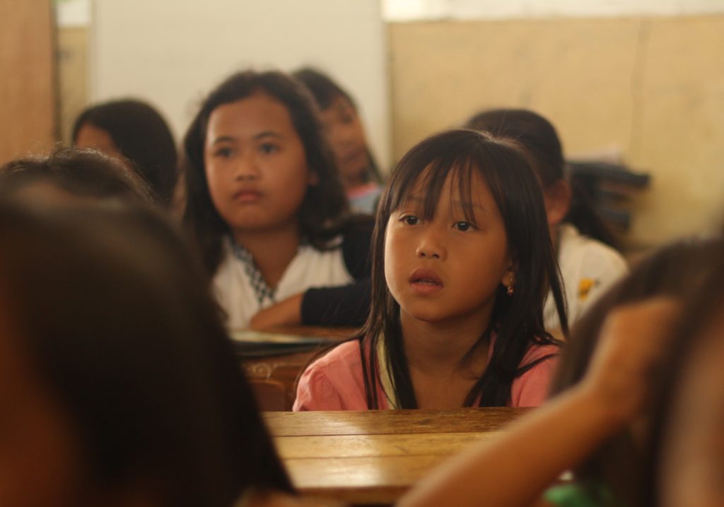 an Asian girl in a classroom full of kids, looking lost at the teacher