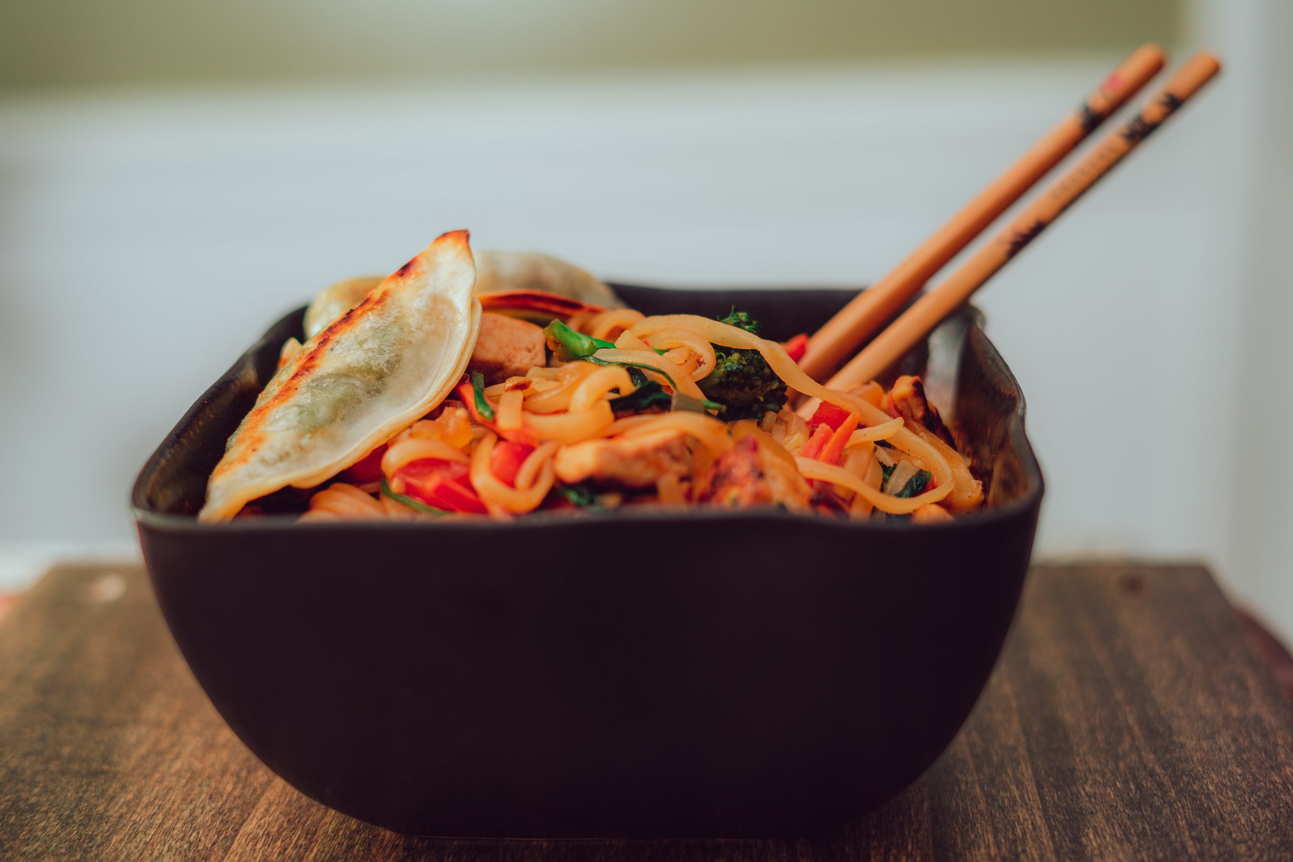 a black bowl with chopsticks filled with chicken pad thai on a brown tabletop