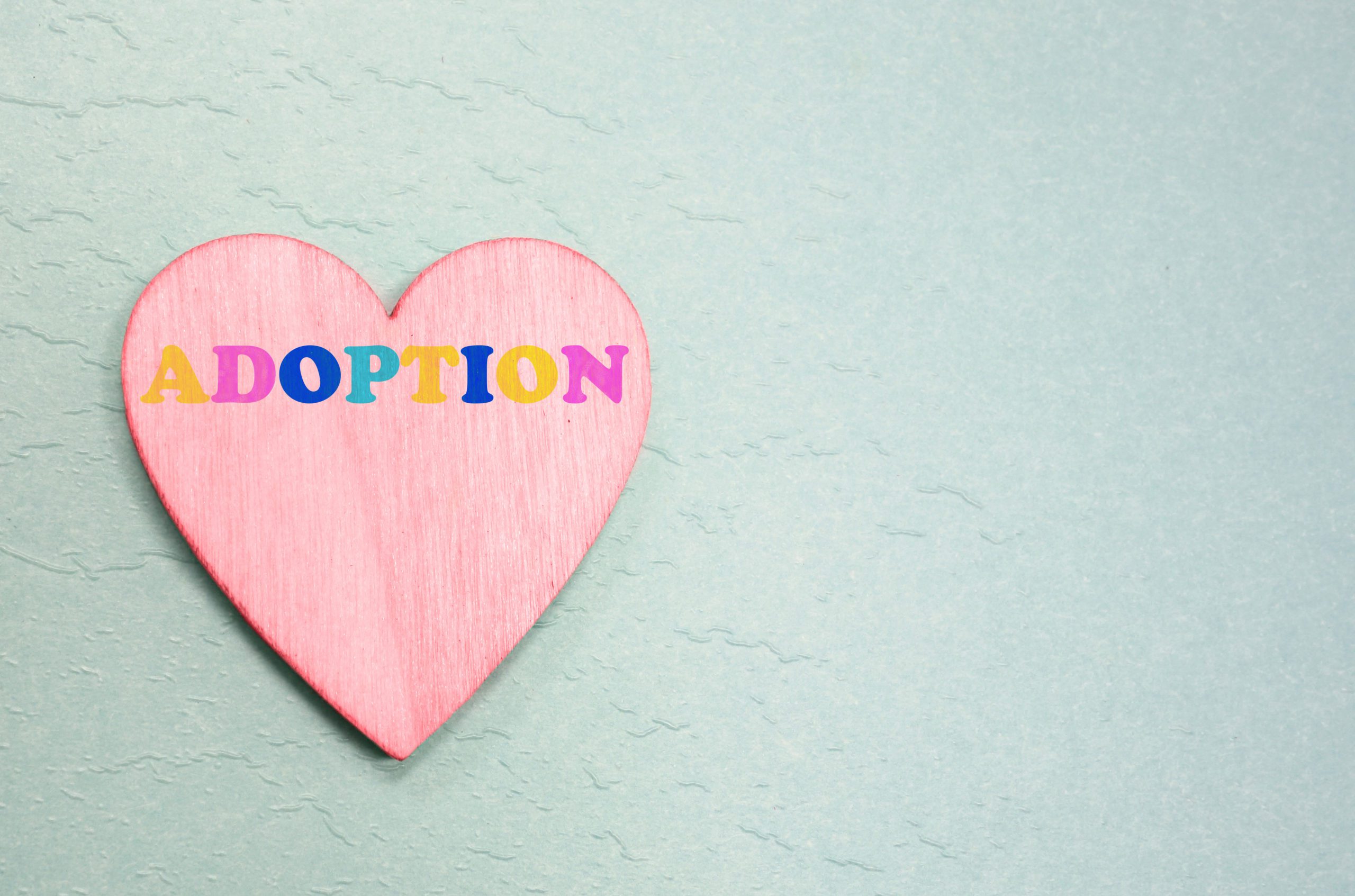 The word adoption written in multicolor letters on a pink heart, with a soft blue background.