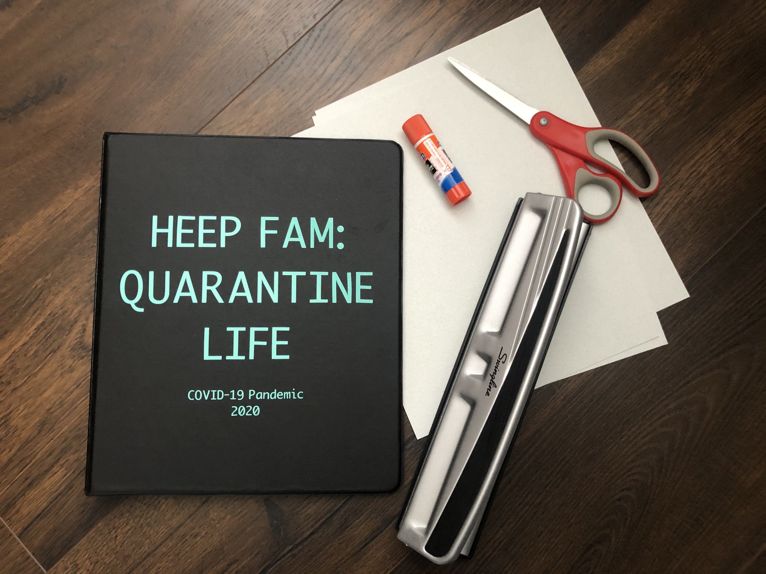 Black quarantine journal with a hole puncher, scissors, and glue stick on a stack of paper on a table