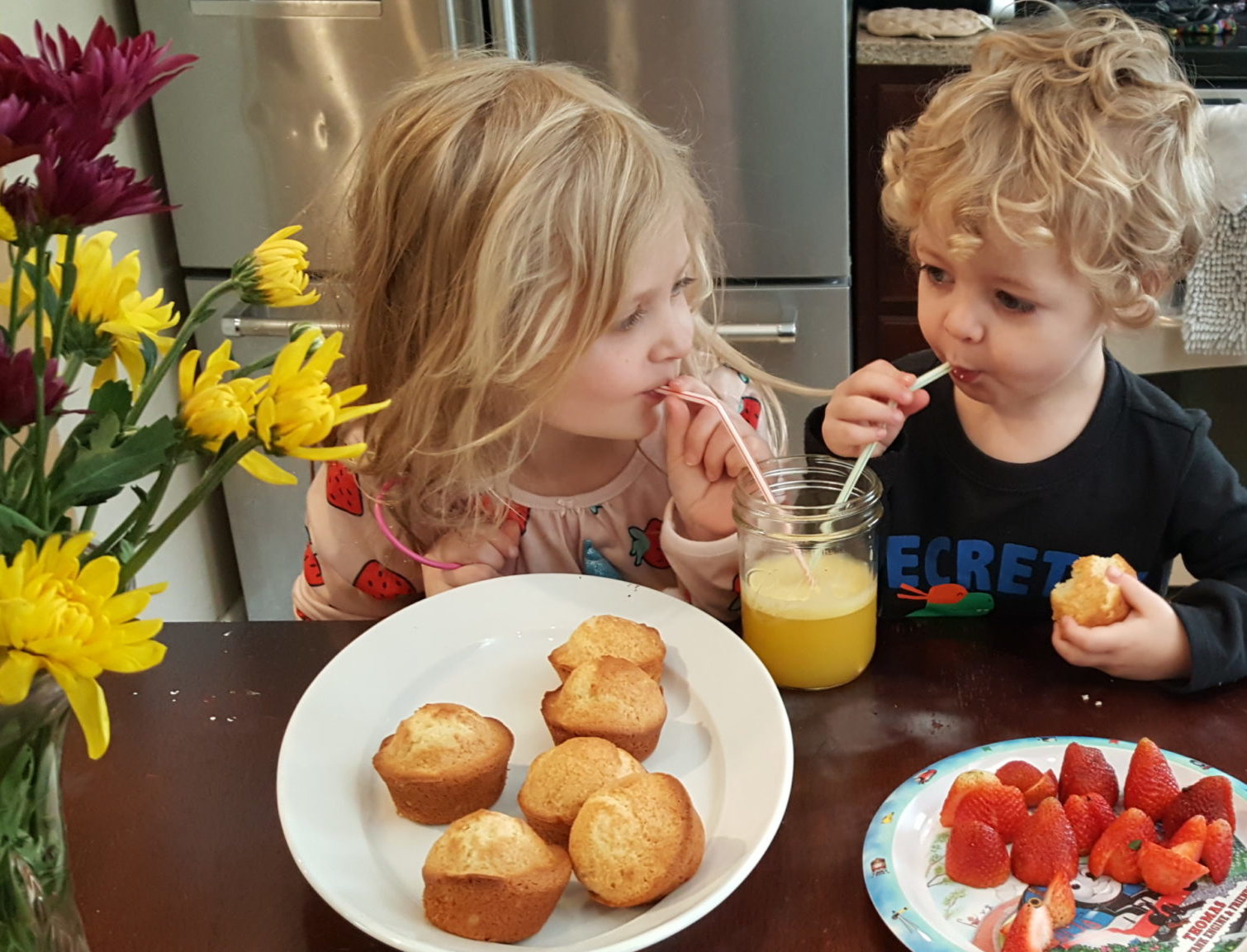 a blonde girl sharing orange juice with two straws with her blonde brother while eating muffins and strawberries