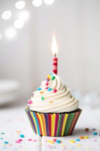 cupcake in a rainbow colored wrapper, with colorful sprinkles and a candle set against a white background