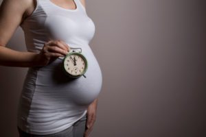 close up of a pregnant woman's belly as she holds a clock up to symbolize her due date