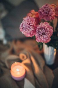a lighted candle on a table next to a white vase of pink flowers as a self-care during isolation strategy