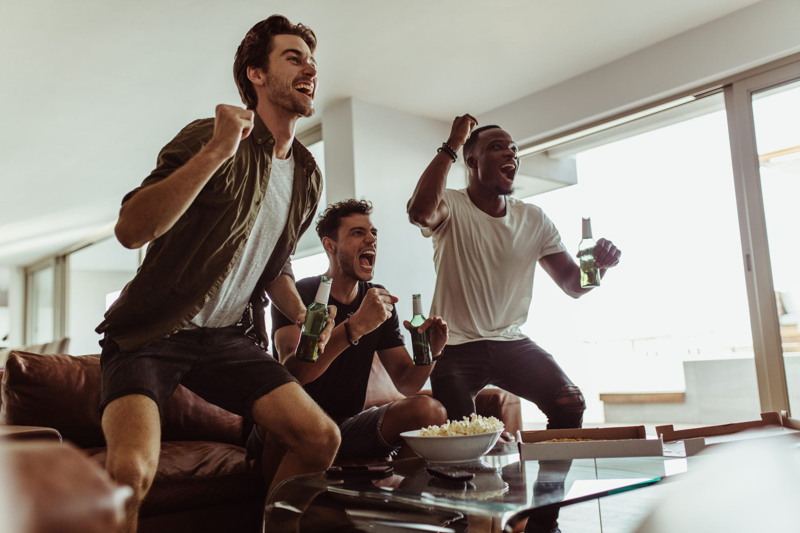 three male friends drinking beer and watching sports on the couch together as they cheer