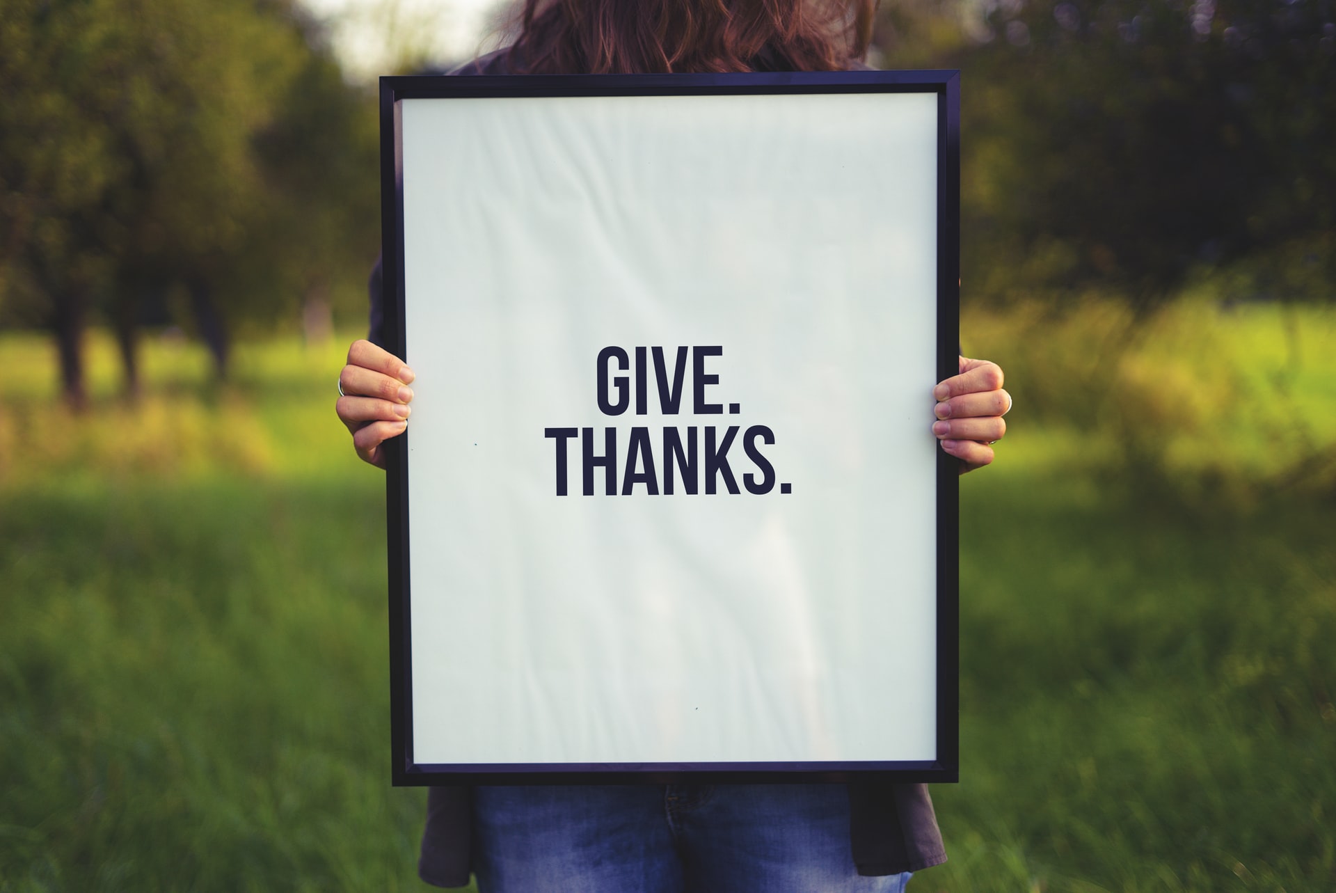 a person showing gratitude by holding a white sign saying, "Give Thanks." in a grassy field.