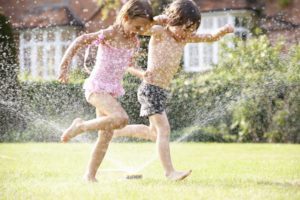two children running through a garden sprinkler, one of summer's best backyard activities