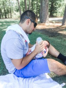 a dad sits on the grass as he bottle-feeds his newborn, symbolic of the support partners give their spouses suffering from postpartum depression