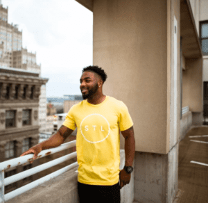 An African American man standing on a rooftop wearing a yellow STL T-shirt