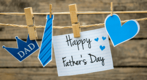A clothesline with a clothespin holding a Happy Father's Day sign with a blue heart, a blue tie, and a crown that says Dad against a wooden slat background