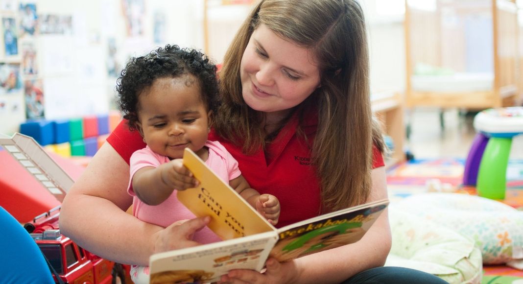 a childcare worker in a red shirt reading a board book to the infant in a pink shirt on her lap
