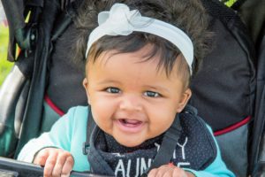 close up of a smiling baby girl with a white flower headband sitting up in a stroller