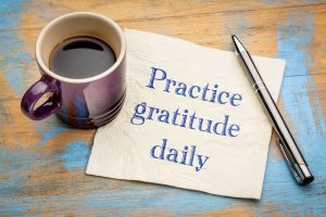 a napkin with the words, "practice gratitude daily? on a wooden tabletop with a cup of coffee and a pen