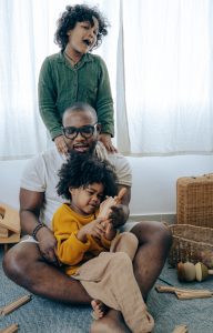 An African American dad playing, with a child on his lap and one on his shoulders