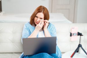 a woman with a laptop on her lap, sitting on a couch as she wipes her tears during an online therapy session as she learns to take care of herself