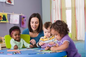 a teacher and three toddlers sitting a table coloring in daycare