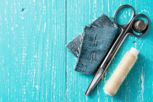 a pair of scissors, a thimble, white thread, and a few needles poked through a square of denim on a distressed turquoise wooden background