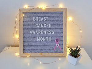 a sign, draped in fairy lights on a white table top that says, "Breast Cancer Awareness Month" with a pink ribbon on it