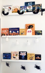 three white wall bookshelves filled with Halloween books, with bats, ghosts, and spiders decorating the shelves