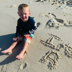 A toddler boy sitting on wet sand with the words, "It's A" spelled out, as they reveal their new baby's gender