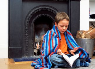 a boy wrapped in a blanket sitting in front of the fireplace with a book
