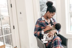 An African American woman sitting in a chair by the window as she breastfeeds her baby