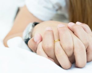 a close up of two people holding hands, one is wearing a wedding ring, as they lay in bed together