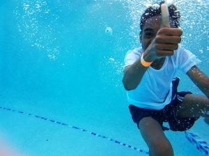 an African American boy swimming underwater as he gives a thumbs up to the camera