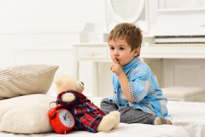 a toddler holding his finger to his lip to shush while he sits on a bed with a teddy bear and alarm clock