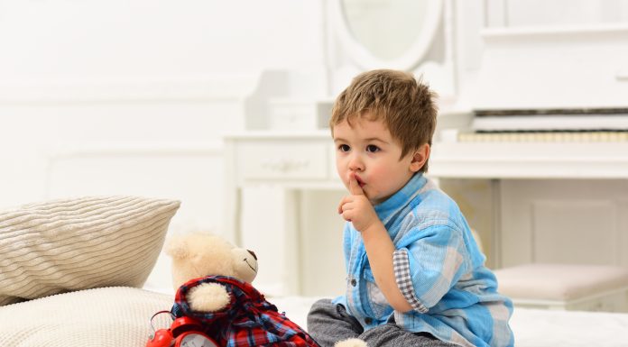 a toddler holding his finger to his lip to shush while he sits on a bed with a teddy bear and alarm clock