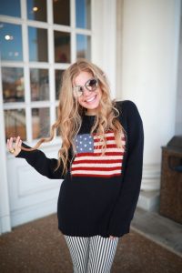 a woman wearing an American flag on her black sweater standing in front of a window