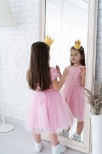 a young girl in a pink dress wearing a gold crown as she looks at herself in a full length mirror
