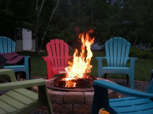 a roaring fire in a fire pit surrounded by empty, multicolored Adirondack chairs