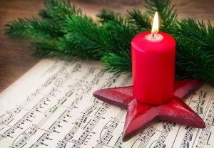 a score of Christmas music on a wooden table with a red, lit candle on top next to a sprig of greenery