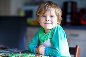 a blonde toddler boy sitting at a table, smiling at the camera as he plays a board game