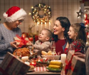 a mom with two girls on her lap as they sit at a Christmas dinner table, with a grandma giving a gift to one of the girls