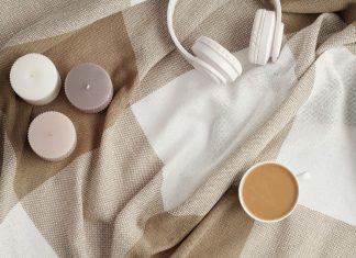 three scented candles, a pair of white headphones, and a cup of coffee on a tan and cream colored blanket