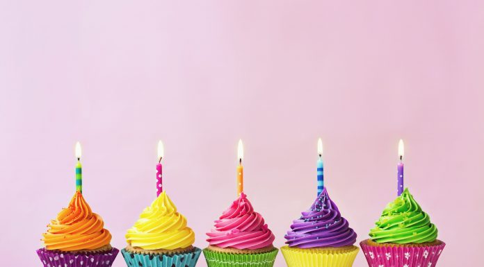 five birthday cupcakes with colorful wrappers and bright colored icing with lit candles on top against a pink background
