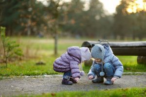 two little girls, bundled up in coats and hats as they squat down on a sidewalk to look at something on the ground