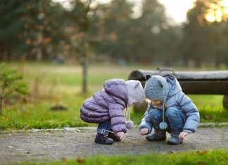 two little girls, bundled up in coats and hats as they squat down on a sidewalk to look at something on the ground