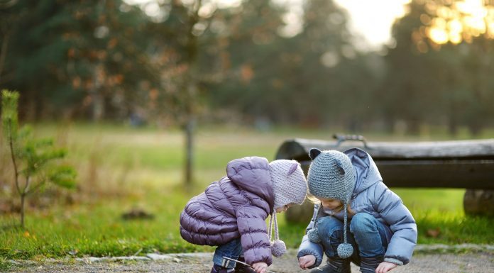 two little girls, bundled up in coats and hats as they squat down on a sidewalk to look at something on the ground