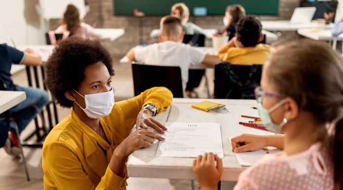 a teacher kneeling by the desk of a student, wearing masks