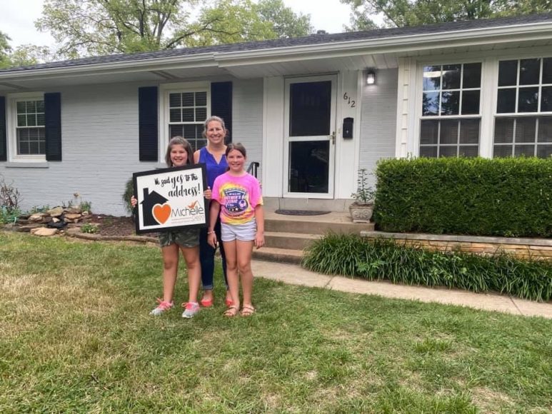 a mom and her two girls standing in front of their new house, holding a sign