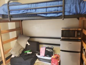 a college dorm room with a loft bed over a mini refrigerator and microwave, and a chair