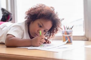 an African American toddler girl laying down and drawing by a window