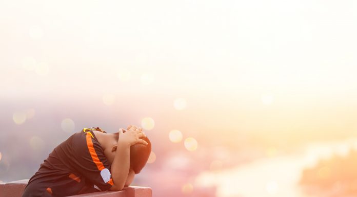 a young boy cradling his head in his hands as he stands outside on a deck overlooking a sun-drenched river