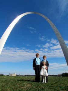 two boys in tuxedos and a flower girl standing under the St. Louis Arch