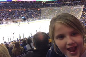 a young girl smiling at the camera with the St. Louis Blues hockey rink behind her