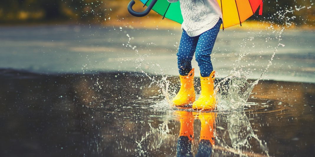 a child in yellow rain boots splashing in puddles as she carries a rainbow umbrella