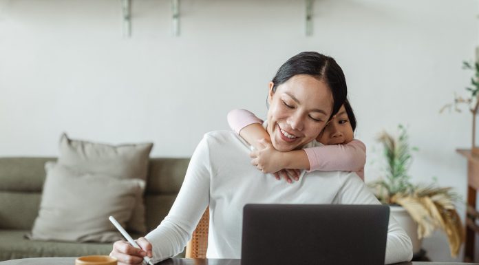 a woman sitting at a desk in front of her open laptop, pen in hand as her toddler daughter wraps her arms around her mom's neck from behind